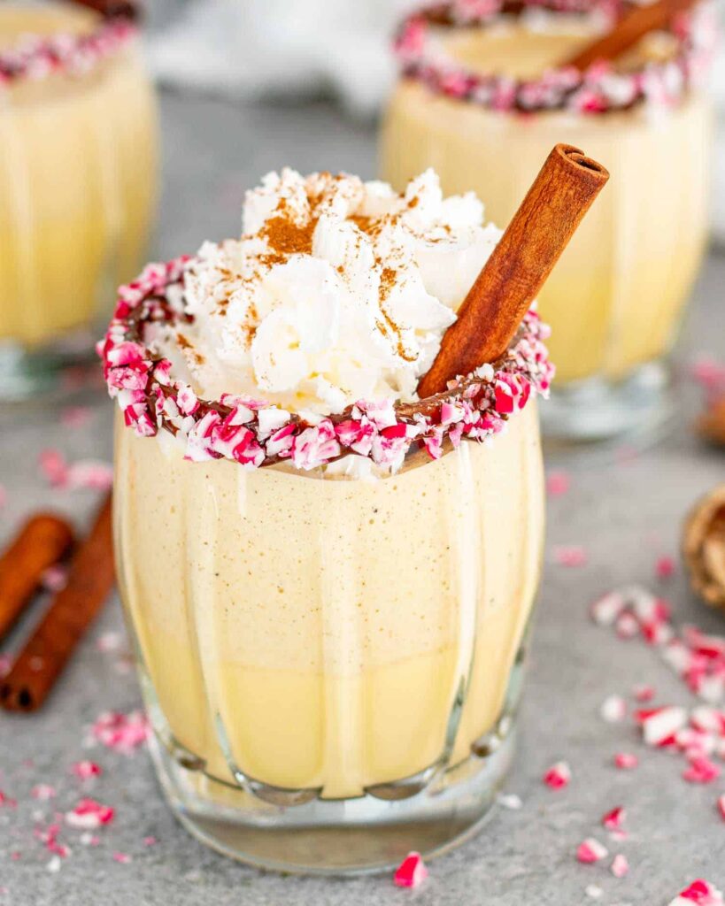 Glasses Of Eggnog In Glasses Rimmed With Chocolate And Crushed Candy Cane Pieces, Garnished With Cinnamon Sticks.